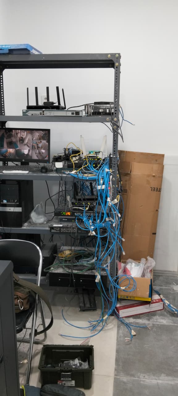 Rak baja terbuka dengan patch cord menggantung sampai ke lantai, dipindahkan ke kabinet 19 inci dengan jalur kabel yang ditata ulang. — Sebelum